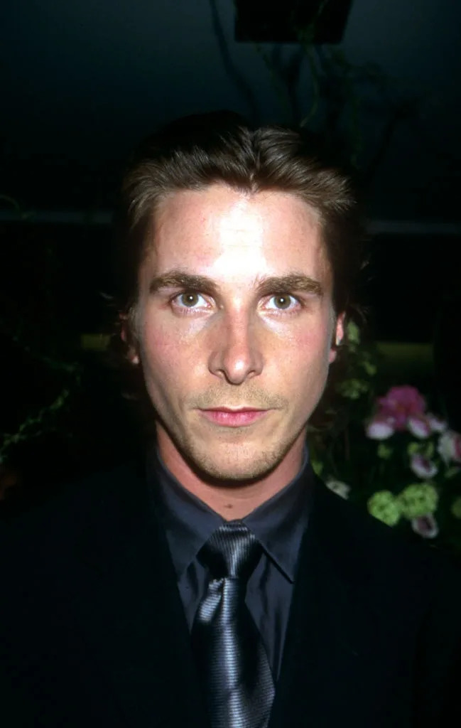 A person in a dark suit and tie at an event, standing indoors with floral decor in the background