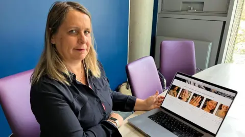 Leanne Rinne/BBC A woman in a dark shirt sat at a table on a purple chair. On the table is an open silver laptop displaying images from the Scarlett Jewels website, likely generated by AI.