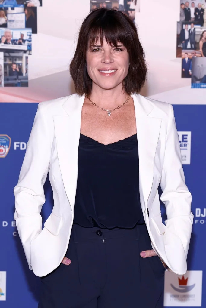 Person in a white blazer and dark outfit on a red carpet, smiling with hands in pockets. Background has posters and logos