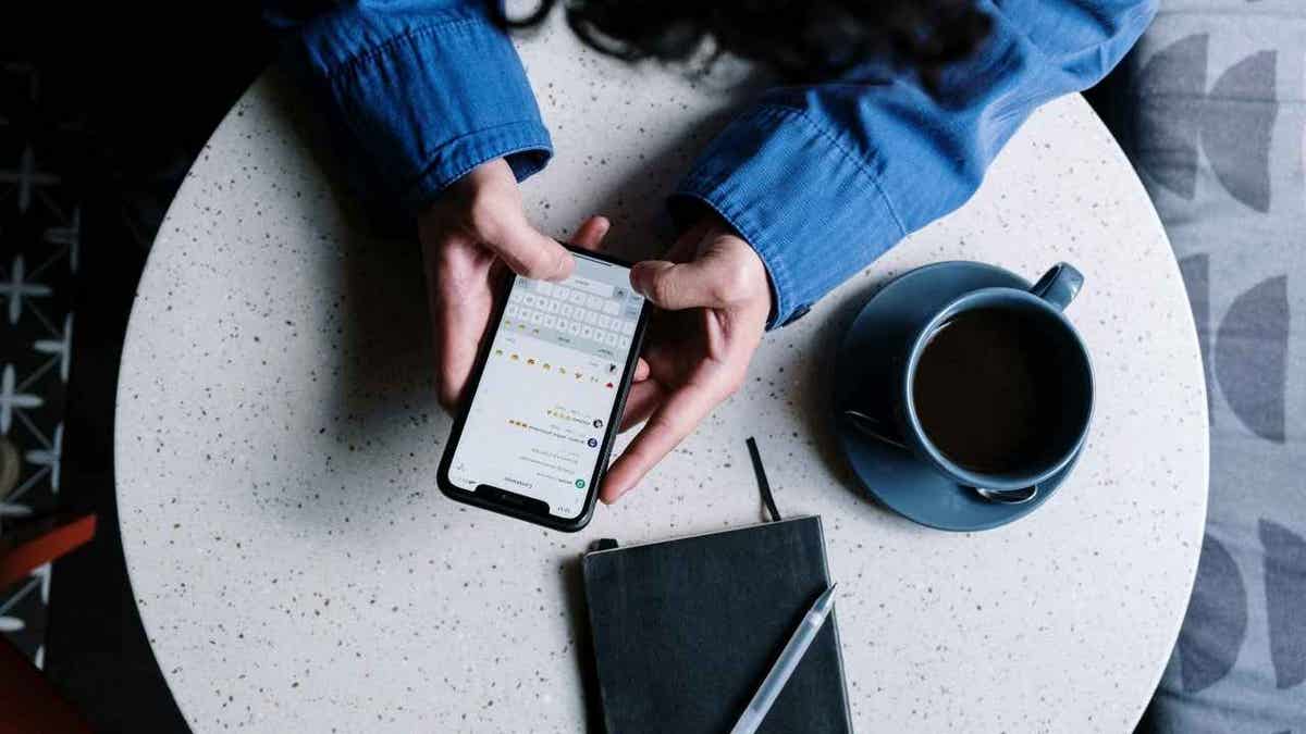 A person wearing a blue shirt uses a smartphone at a round table with a notebook, pen, and cup of coffee.