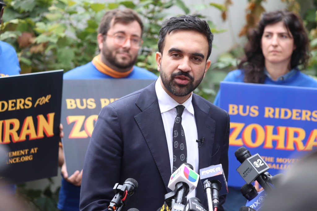 2025 NYC Mayoral candidate Zohran Mamdani speaking at a press conference with microphones from various news outlets.