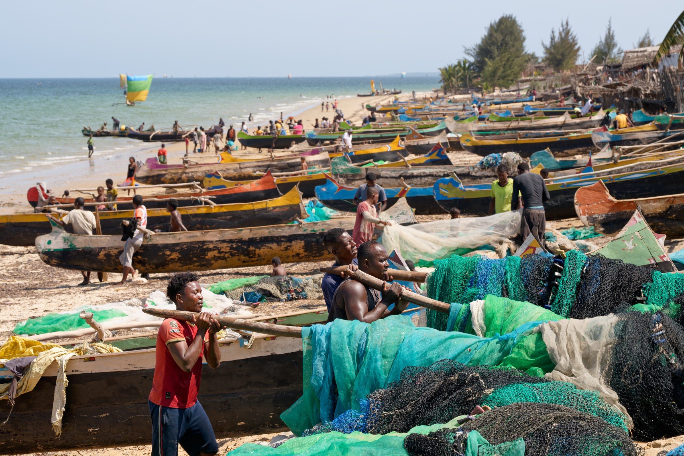 Fishermen return to a beach crowded with boats, the men carrying heavy fishing gear after a morning working at sea.