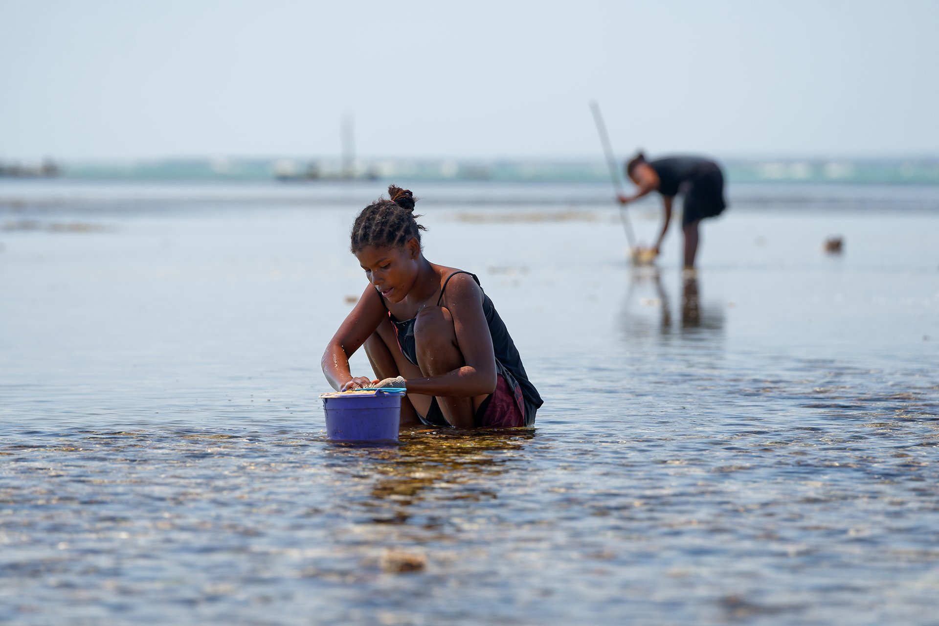A woman crouched in the sea with a blue bucket.