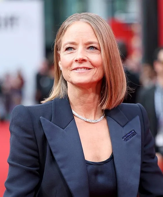 A person wearing a dark, tailored suit with a necklace on a red carpet, posing for photos