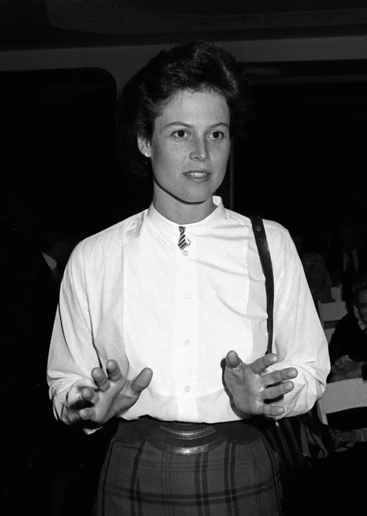 Person in a button-up shirt and plaid skirt gestures while speaking at an event