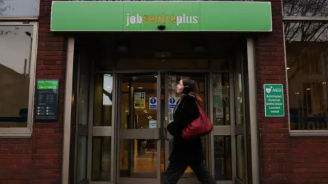 EPA A stock image shows a young woman walking past the entrance to a Job Centre plus. She is wearing headphones and carrying a red bag. Above her, the words job centre plus are written in white and yellow against a green background.