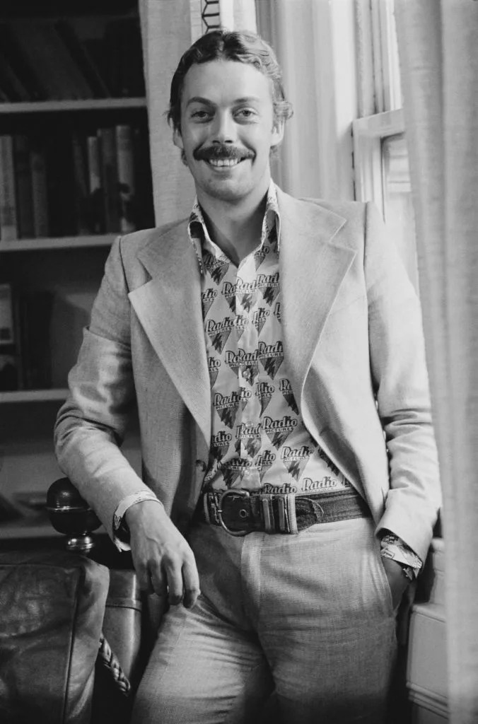 Man in a patterned shirt and tailored blazer, leaning casually by a window