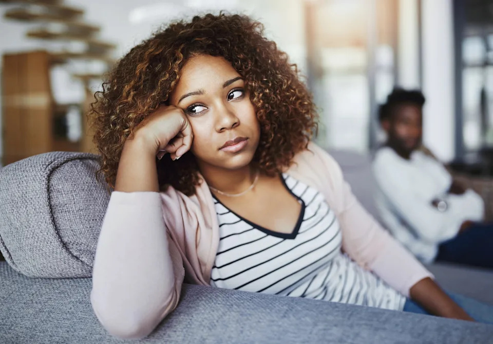 Woman with curly hair looks contemplative on a couch, wearing a striped shirt and cardigan, with a blurred person in the background