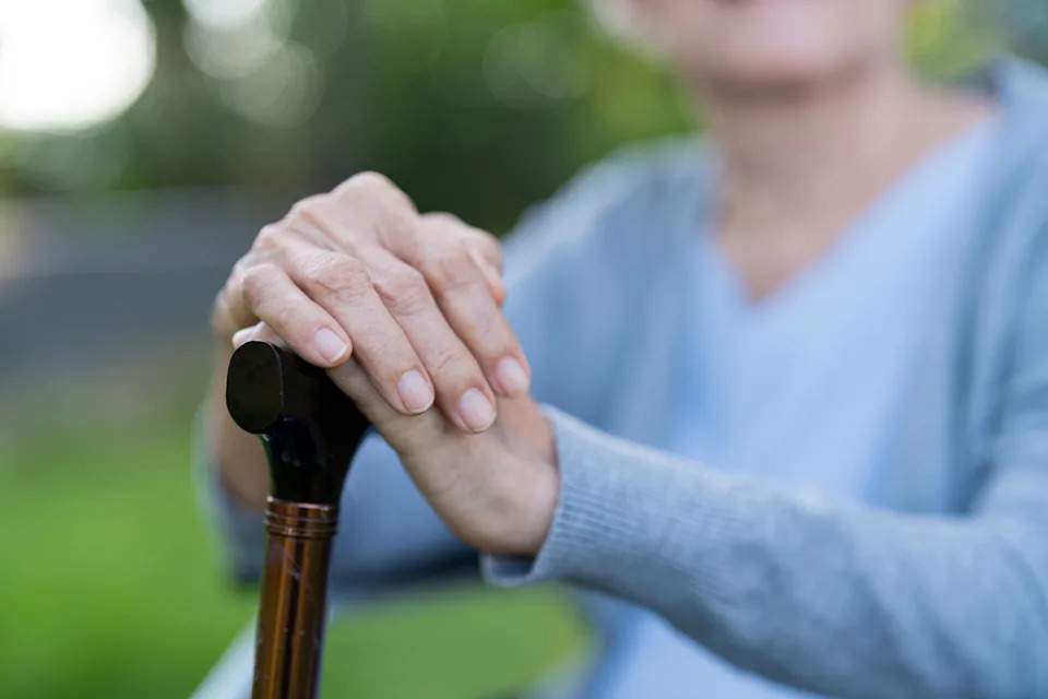Elderly person resting hands on a walking cane, sitting outdoors, wearing a sweater