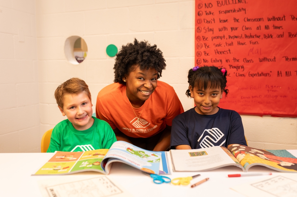Two smiling children and one staff member reading books at the Boys & Girls Club