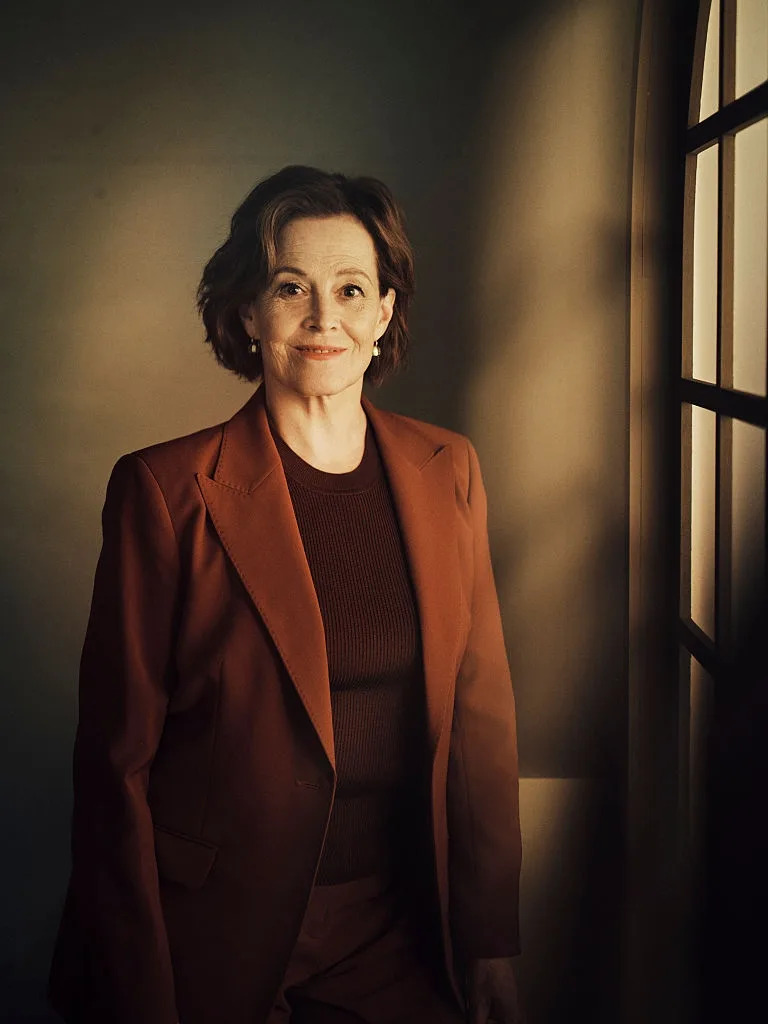 Actress in a tailored suit stands by a window, smiling subtly, blending classic and sophisticated style