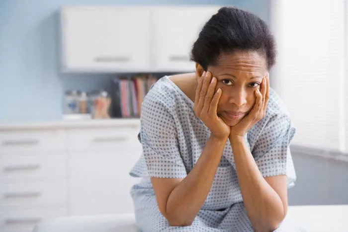 A concerned person in a hospital gown sits in a medical setting, resting their face in their hands, suggesting contemplation or worry
