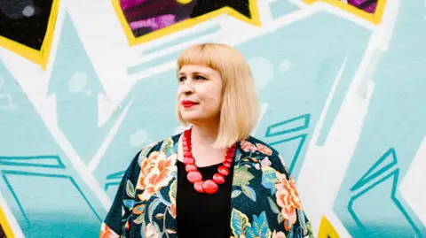 Jess Lowe A woman with blonde hair and a floral top is standing in the centre of frame in front if a blue wall with a funky pattern.