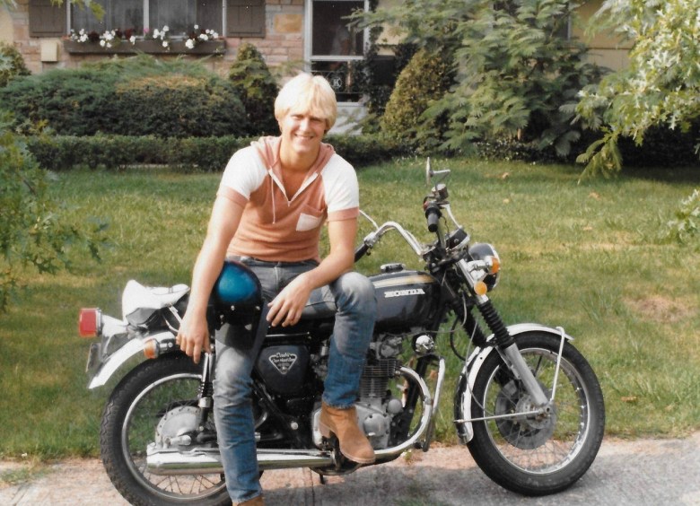 An aging photo of a man sitting on the side of a motorcycle parked on a cement drive in front of a partly brown lawn.