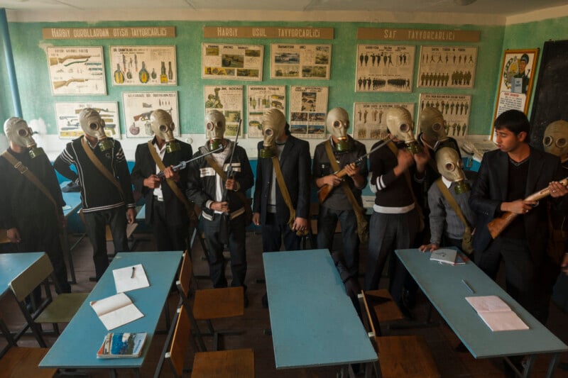 Students in a classroom stand in a row wearing gas masks and holding rifles, with educational posters on the walls and notebooks on empty blue desks.