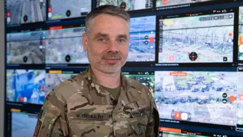 BBC/Matthew Goddard Yuriy Myronenko stands in army fatigues in front of a wall of screens showing fields, scrublands and some houses. All scenes from the Russian-Ukrainian frontline
