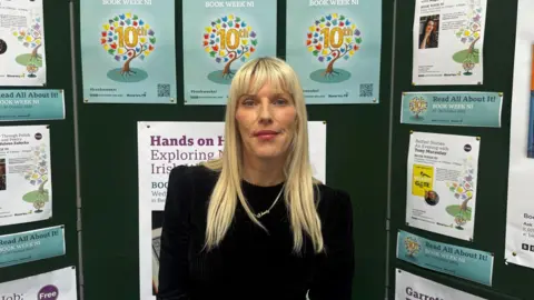 BBC Wendy Erskine has long blonde hair wearing a black dress is smiling at the camera. There are posters for Book Week NI in the background. 