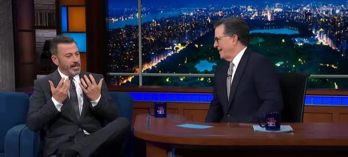Two men in suits talk on a late-night talk show set, with a cityscape backdrop behind them
