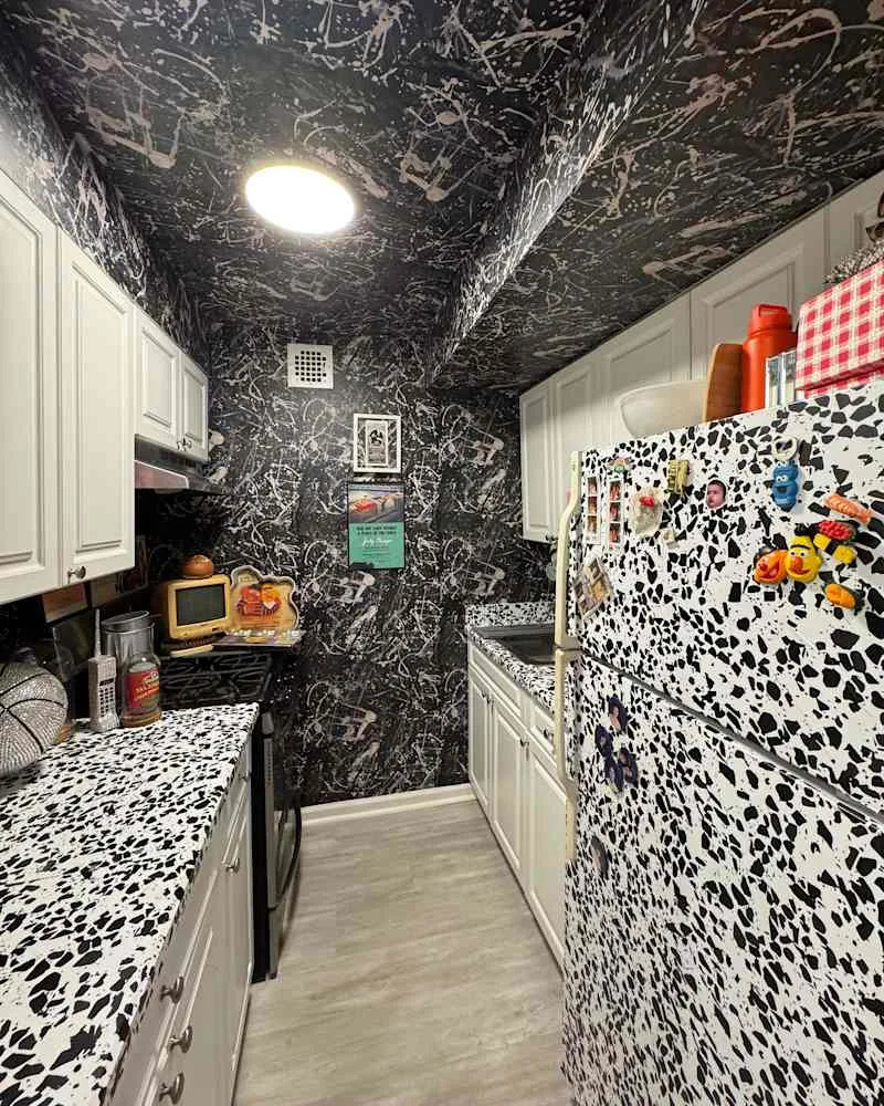 Narrow kitchen with black and white splatter wallpaper, white cabinets, and a patterned countertop.