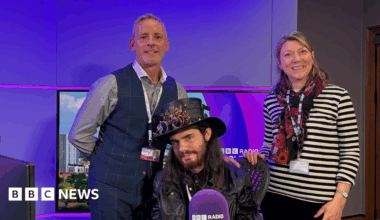 Rob Amey and Radio Solent presenter Katie Martin standing behind Logan sitting in the radio studio chair. The studio lights are purple. Logan is wearing a steam punk style top hat with elaborately decorated goggles resting on the brim. He has long dark hair and a full beard and is wearing a leather biker jacket. Rob Amey has short fair hair and is wearing a grey long-sleeve shirt and a royal blue waistcoat. Katie Martin has shoulder-length blonde hair and is wearing a black and white horizontal striped top and a multi-colour scarf.