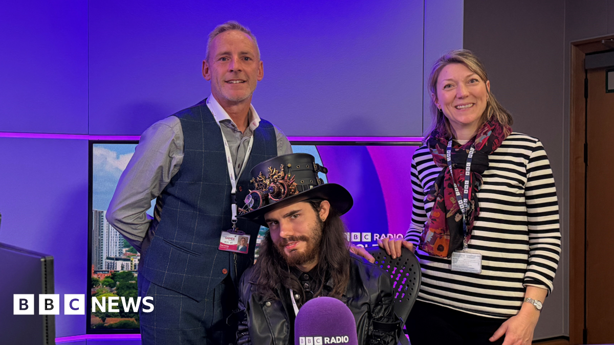 Rob Amey and Radio Solent presenter Katie Martin standing behind Logan sitting in the radio studio chair. The studio lights are purple. Logan is wearing a steam punk style top hat with elaborately decorated goggles resting on the brim. He has long dark hair and a full beard and is wearing a leather biker jacket. Rob Amey has short fair hair and is wearing a grey long-sleeve shirt and a royal blue waistcoat. Katie Martin has shoulder-length blonde hair and is wearing a black and white horizontal striped top and a multi-colour scarf.