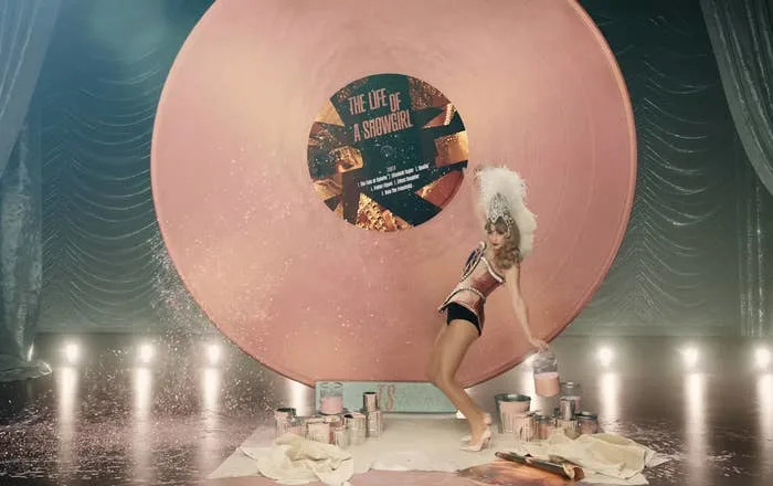 A performer poses in front of a giant pink vinyl record prop, wearing a flamboyant headdress and bodysuit, surrounded by paint cans