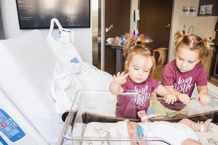 Two young girls in "big sister" shirts curiously look at a newborn baby in a hospital bassinet