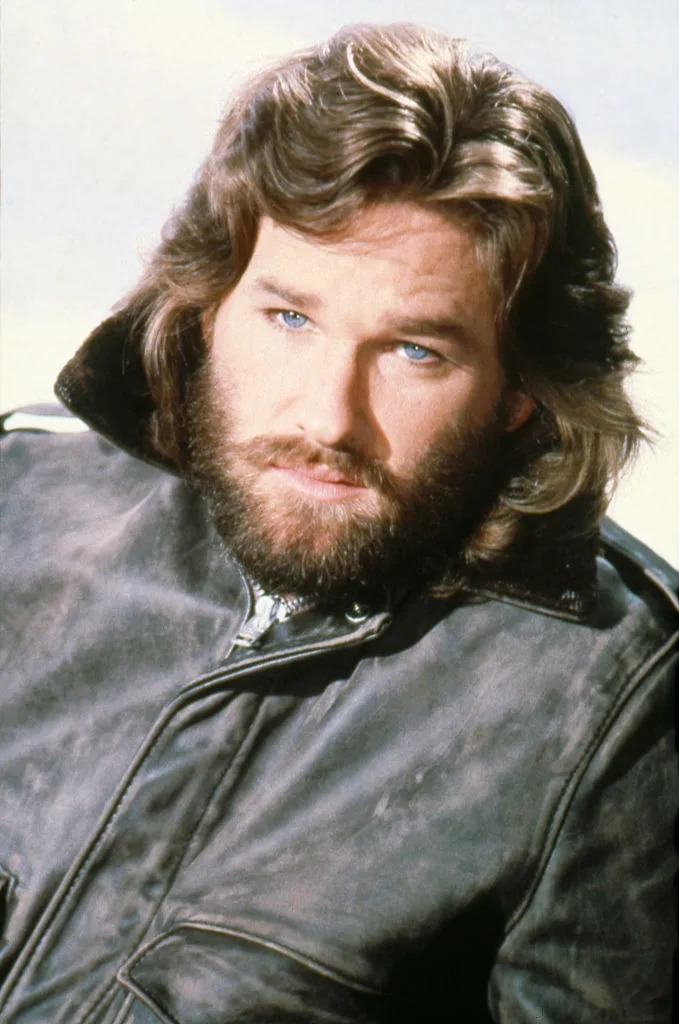 Man with wavy hair and a beard, wearing a leather jacket with a classic style, gazing intently ahead