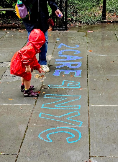 A child in a red raincoat stands over some writing on a sidewalk.
