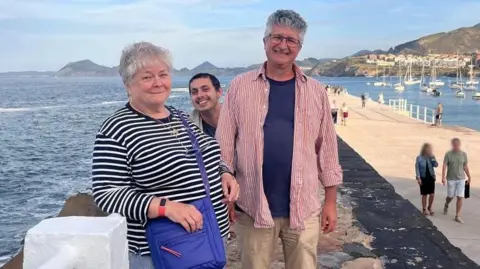 Alan Smith Alan Smith stands with his wife and their daughter's boyfriend on the pier at Castro Urdiales. The pier can be seen stretching out behind them with fishing boats and hills seen in the background.