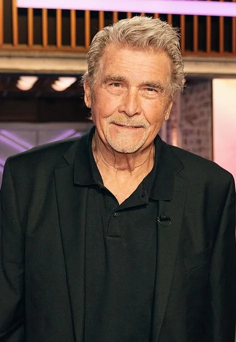 A person in a black suit stands in a studio with "The Kelly Clarkson Show" displayed on a screen in the background