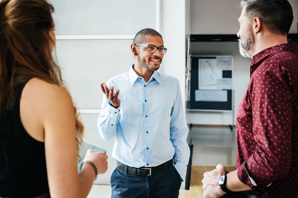 A man in a smart casual outfit is speaking to two people in a professional setting, gesturing with his hand while smiling