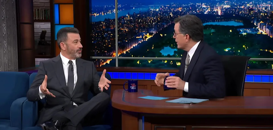 Two talk show hosts in suits have a lively discussion on a TV set with a cityscape background