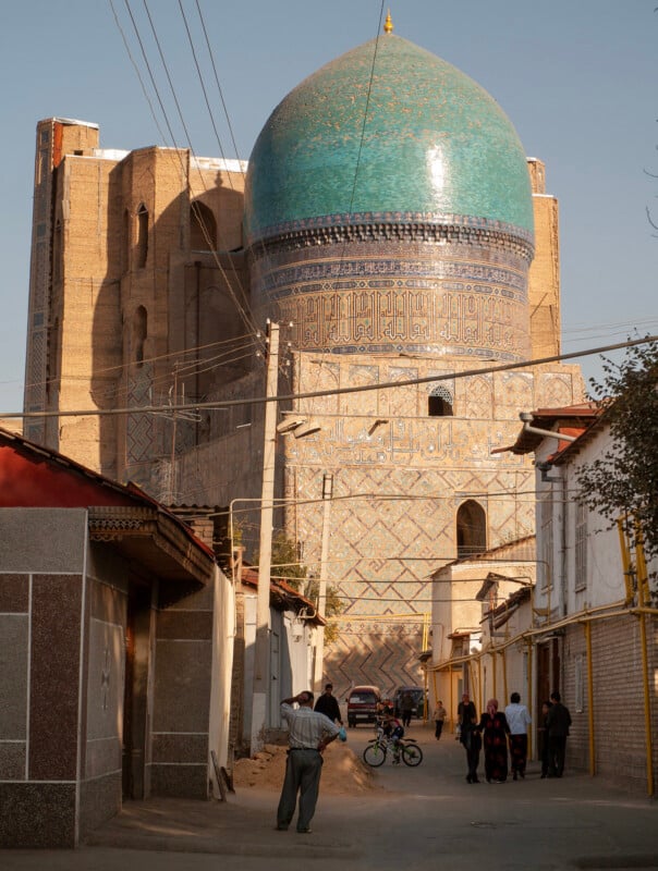 A group of people walk and ride bicycles down a narrow street lined with buildings, with a large historic structure featuring a turquoise dome and intricate tilework towering in the background.