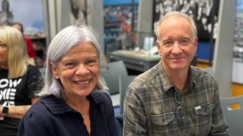 Jane McComb on left smiling at camera. Grey hair and navy jumper. Graham Mounsey sat beside her on the right, also smiling at camera. Wearing a green shirt. 