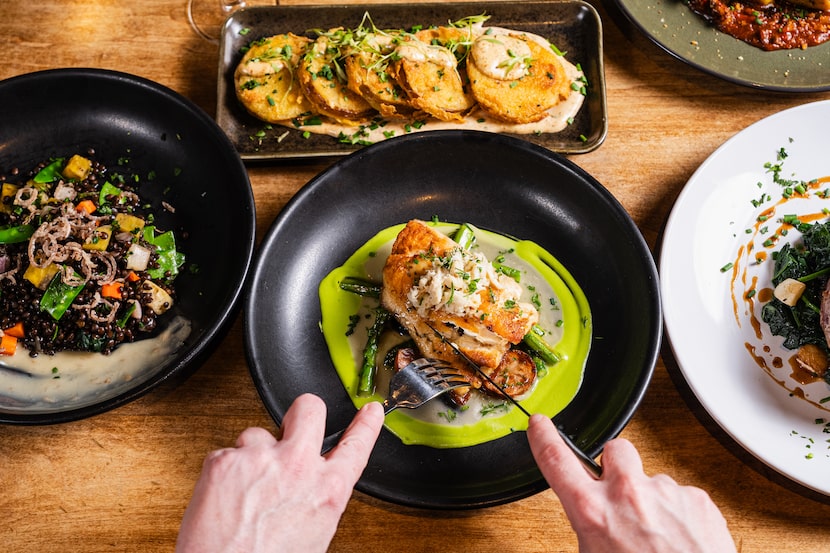 Hands holding silverware slicing into food on a black ceramic plate. Clusters of other...