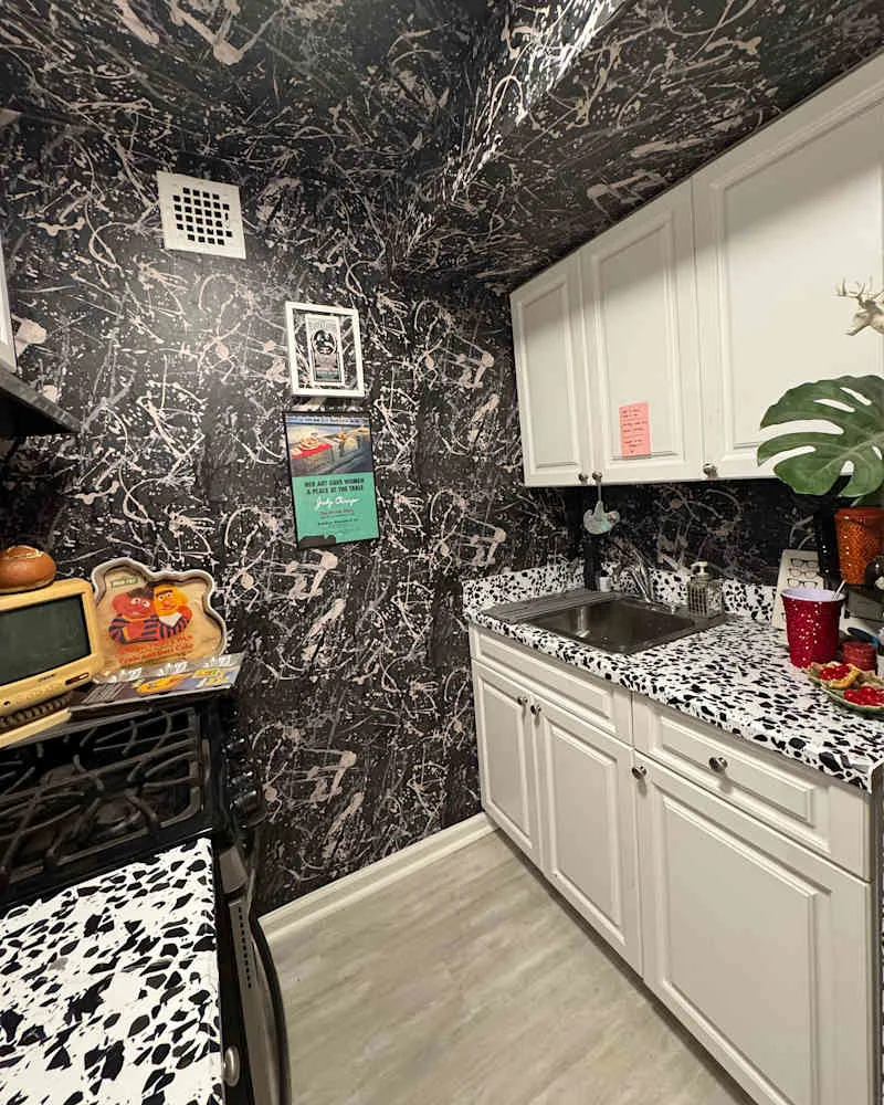 Compact kitchen with black and pink abstract wallpaper, white cabinets, a sink, and a speckled countertop.