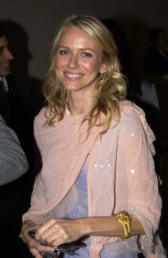 Actress wearing a draped, delicate shawl, holding sunglasses. She is smiling at an event, interacting with others around her