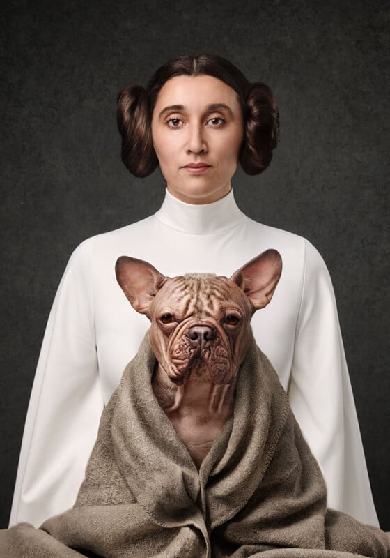 A woman dressed as Princess Leia in a white gown with side buns poses with a wrinkled dog wrapped in a gray blanket, resembling the iconic Star Wars character and her companion, against a dark backdrop.