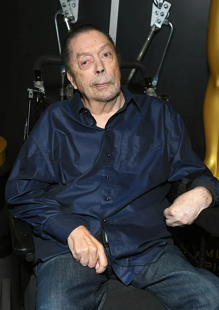 A person in a dark shirt, seated in a wheelchair, poses at an event with an entertainment award statue visible in the background