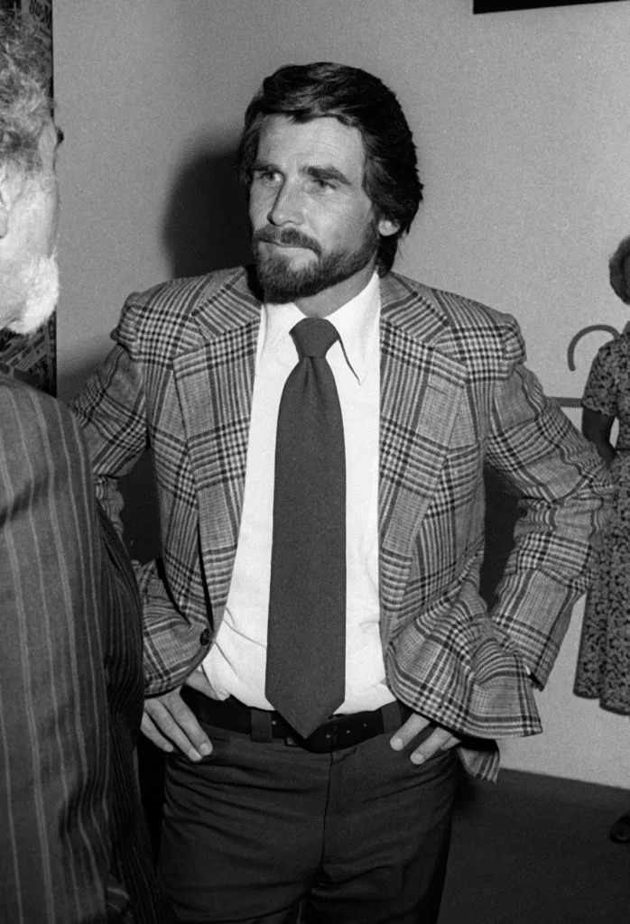 Man in a plaid suit, hands on hips, converses with another person. Vintage setting