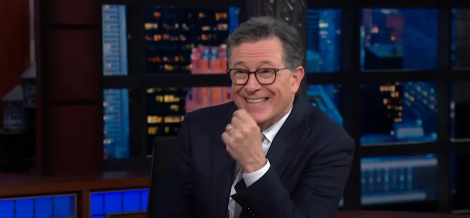 A person in a suit and glasses is smiling and sitting at a nighttime talk show desk against a cityscape backdrop
