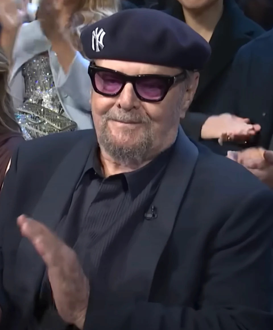 Person in dark suit and glasses, wearing a beret, clapping at an indoor event