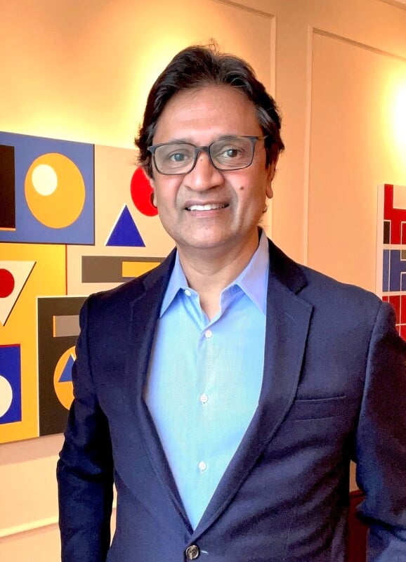 A man wearing glasses, a blue shirt, and a dark blazer stands indoors, smiling in front of a wall decorated with modern, colorful geometric artwork.