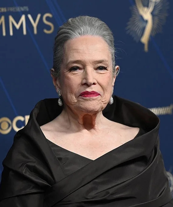 Person in elegant, draped black gown on a red carpet at an Emmy event, with ornate earrings and swept-back hairstyle