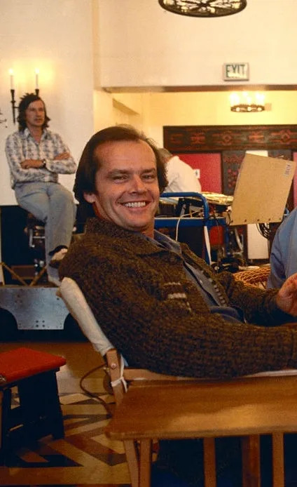 Actor smiling on a film set, sitting on a chair next to a director who is focused on a screen. Production crew and equipment visible in the background