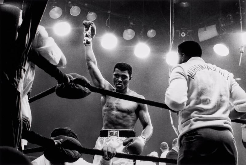 A victorious boxer stands in a ring with one glove raised high. Bright lights shine overhead, and team members in jackets, one labeled "Muhammad Ali," stand nearby. The crowd and other figures are partially visible.