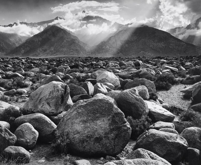 Large boulders scattered across a rocky plain with dramatic mountains in the background, partly shrouded by clouds and rays of sunlight breaking through. The scene is in black and white.