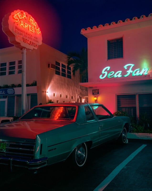 A vintage teal car is parked at night outside the Sea Fan Apartments, illuminated by neon signs and warm lighting. The building and retro signage evoke a nostalgic, mid-century atmosphere.