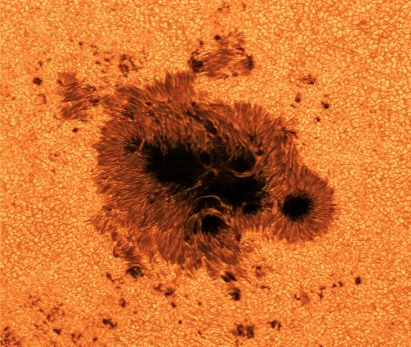 Close-up image of a large sunspot on the Sun’s surface, showing a dark central region surrounded by textured, glowing orange and granular patterns.